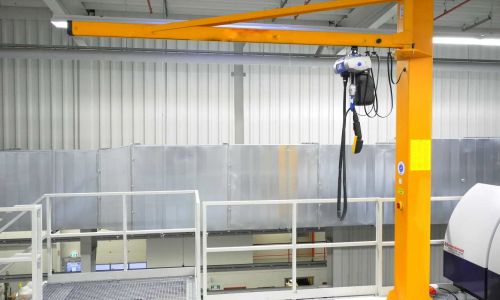 Gelber Schwenkkran mit elektrischem Seilzug in Industriehalle, montiert auf erhöhter Plattform mit Geländer.