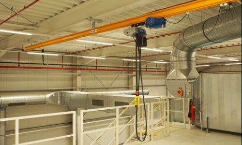 Leichter Deckenkran mit Kettenzug in einer Industriehalle mit Lüftungsrohren und Geländern.
