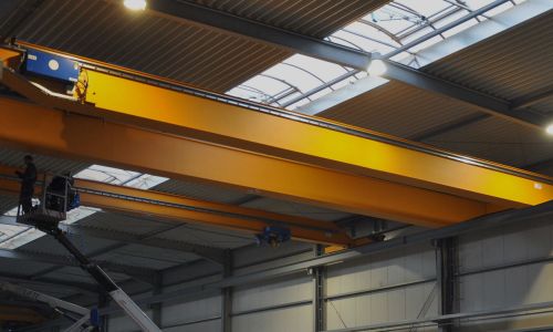 Großer gelber Hallenkran mit Laufkatze und Haken unter Hallendach mit Lichtkuppeln.